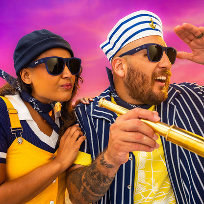 A man and woman in navy blue sunglasses with amber gradient lenses and matching sailor gear watch a beautiful ocean sunset.