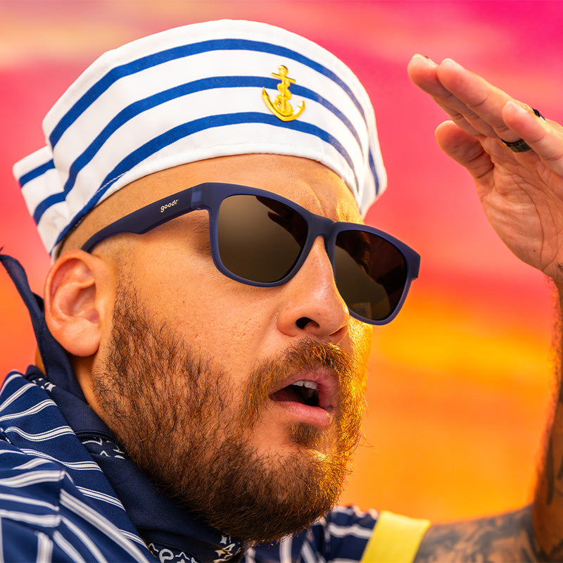 A man in a striped sailor cap and navy blue sunglasses with amber gradient lenses excitedly looks into the horizon at sunset.